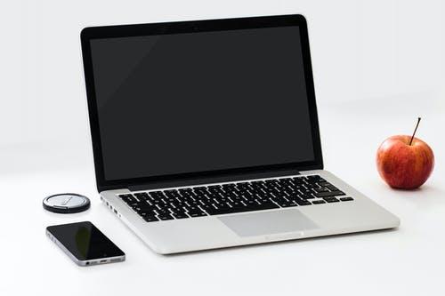 En bärbar dator som ligger uppslagen på ett bord med en mobiltelefon på vänster sida och ett rött äpple på höger sida.
