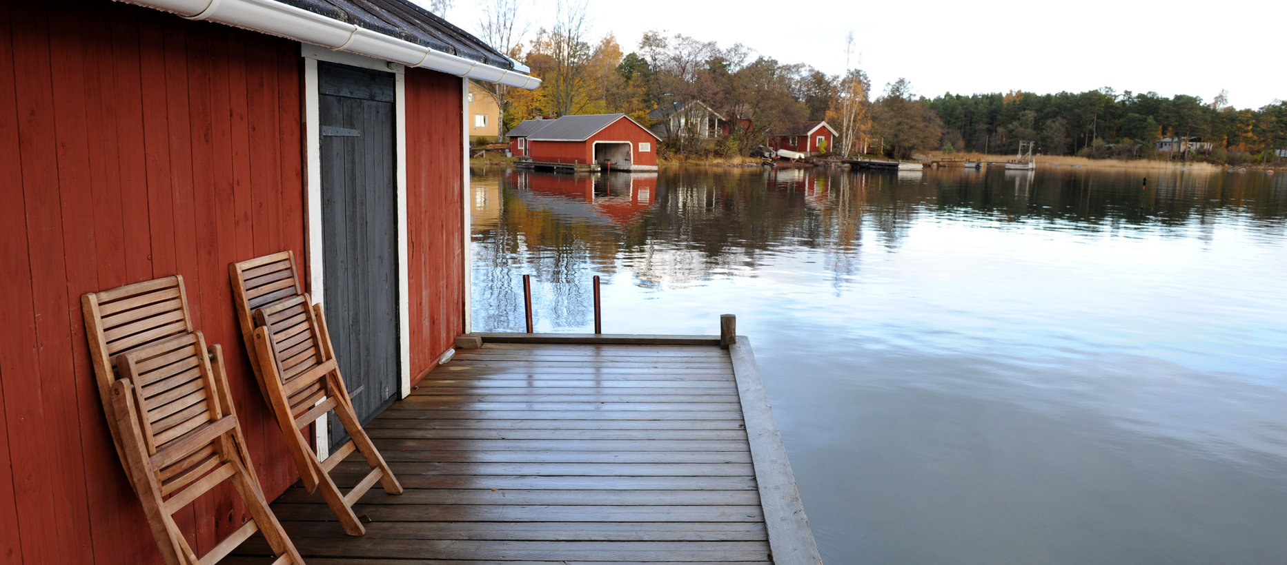Hitis röda båthus med två ihopfälda stolar på bryggan. Vattnet ligger spegelblankt.