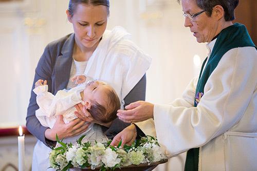 En kvinnlig präst döper ett barn som är i kvinnas famn. Dopfunten är smyckad med vita blommor.